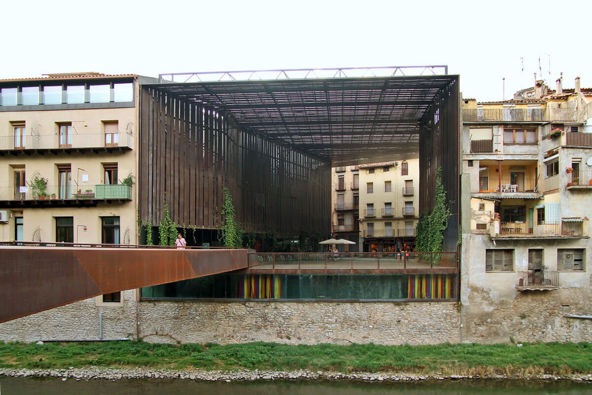 Covered urban space between existing buildings overlooking the Ter river, wrapped with steel slats