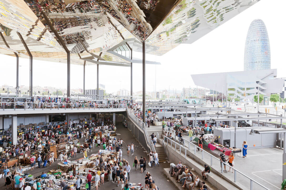 Busy flea market beneath golden canopy with faceted geometry facing Barcelona's Torre Gloriès
